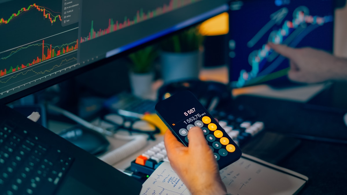 Trader calculating position size with a calculator beside live market charts